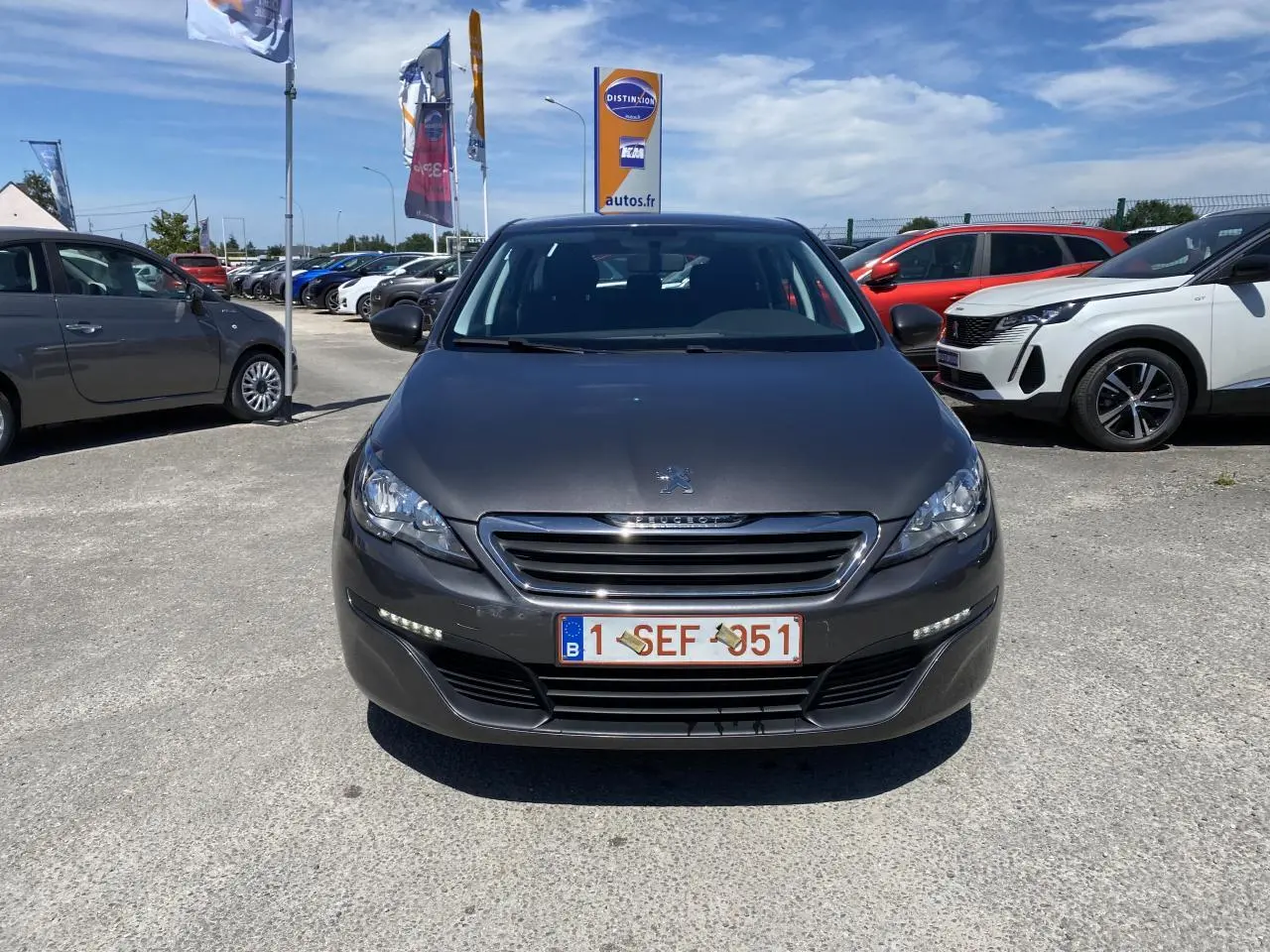 Vue frontale d'une Peugeot 308 SW gris clair garée en extérieur sous un ciel bleu avec d'autres véhicules autour.