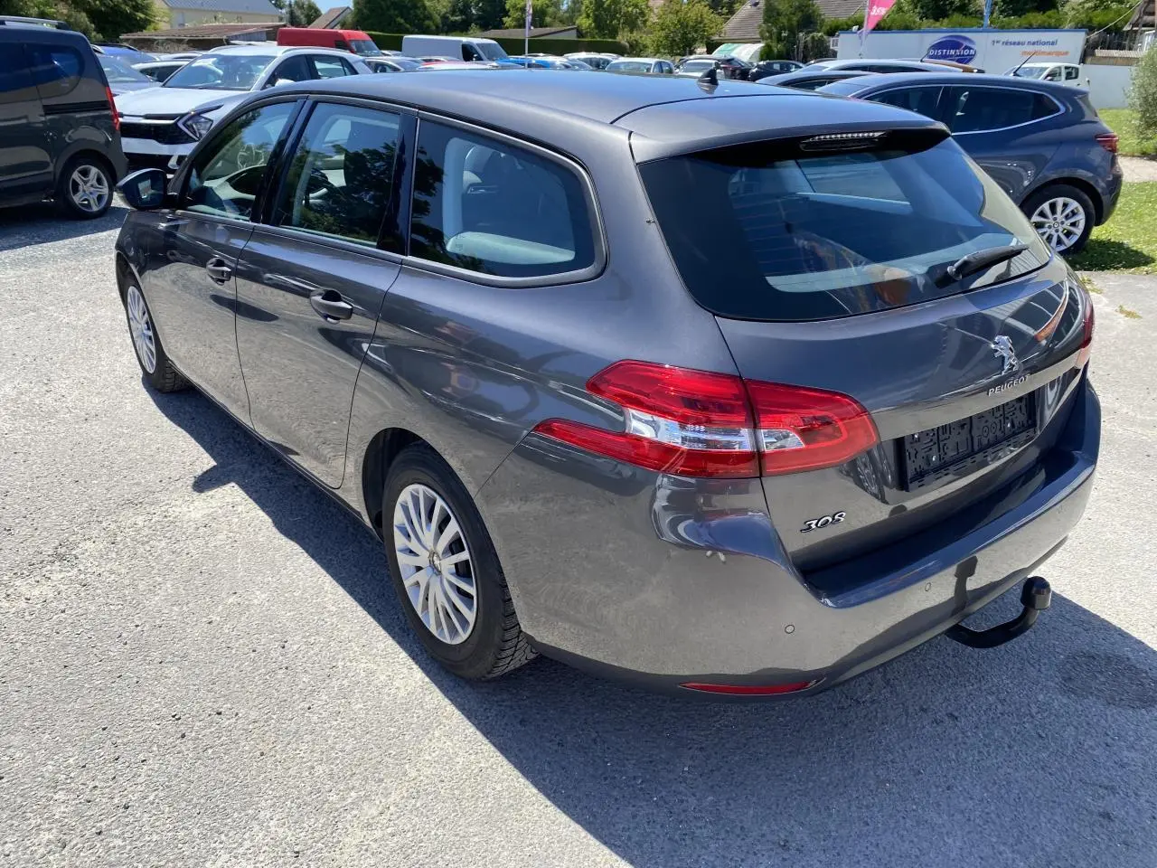 Peugeot 308 SW gris clair vue 3/4 arrière droit avec attelage et feux arrière distinctifs au soleil.
