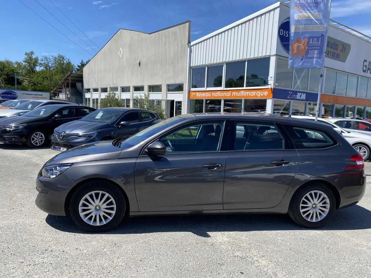 Profil côté gauche d'une Peugeot 308 SW gris clair garée devant un bâtiment commercial sous un ciel bleu.