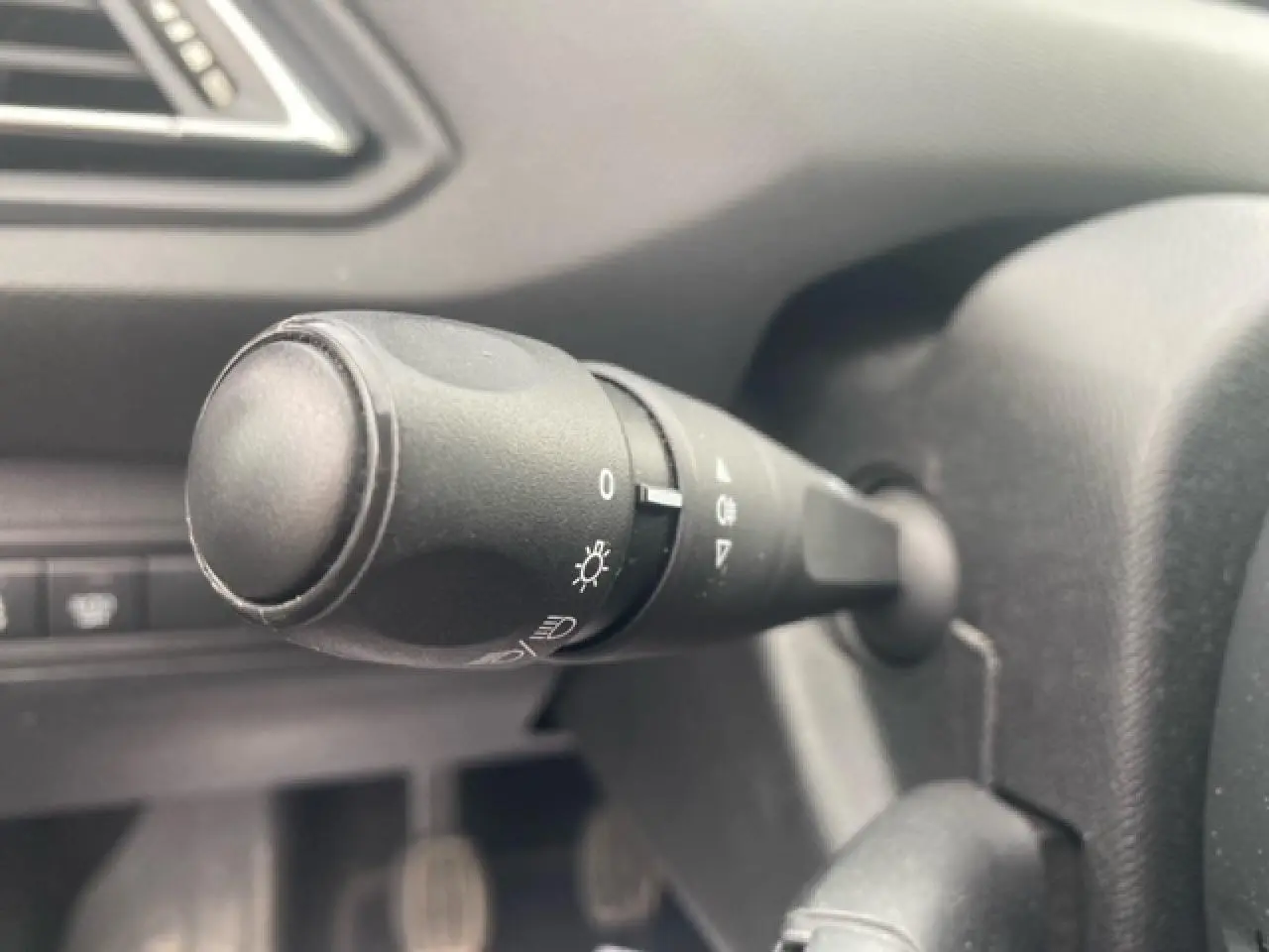 Gros plan sur la manette de commande des phares à gauche du volant dans l'habitacle d'une Peugeot 308 SW gris clair.