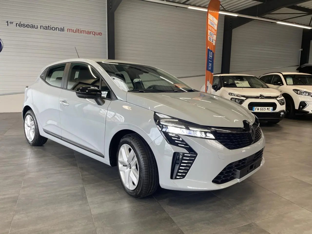 Renault Clio 1.5 Blue dCi 2025 gris Rafale vue 3/4 avant en intérieur avec phares LED et jantes argentées.
