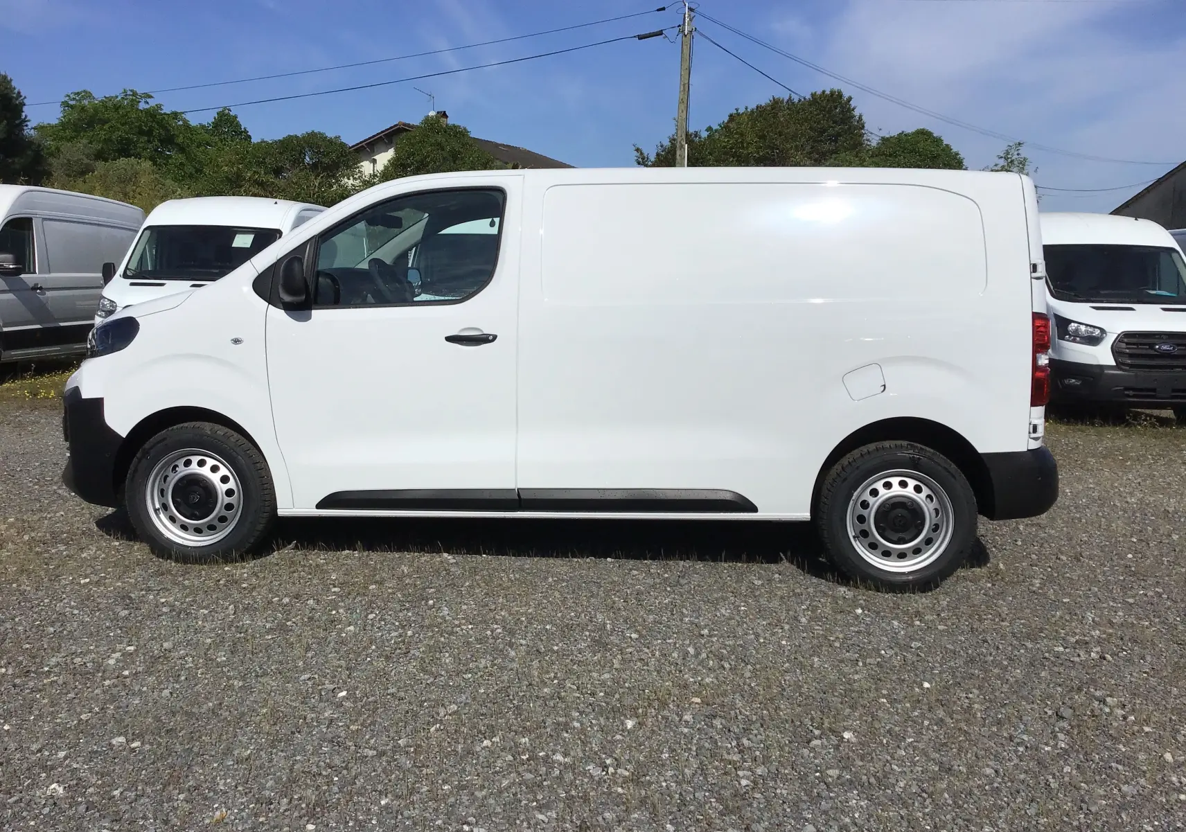 Peugeot Expert Fourgon blanc vu de profil côté gauche, avec carrosserie lisse et roues acier, stationné sur sol gravillonné.