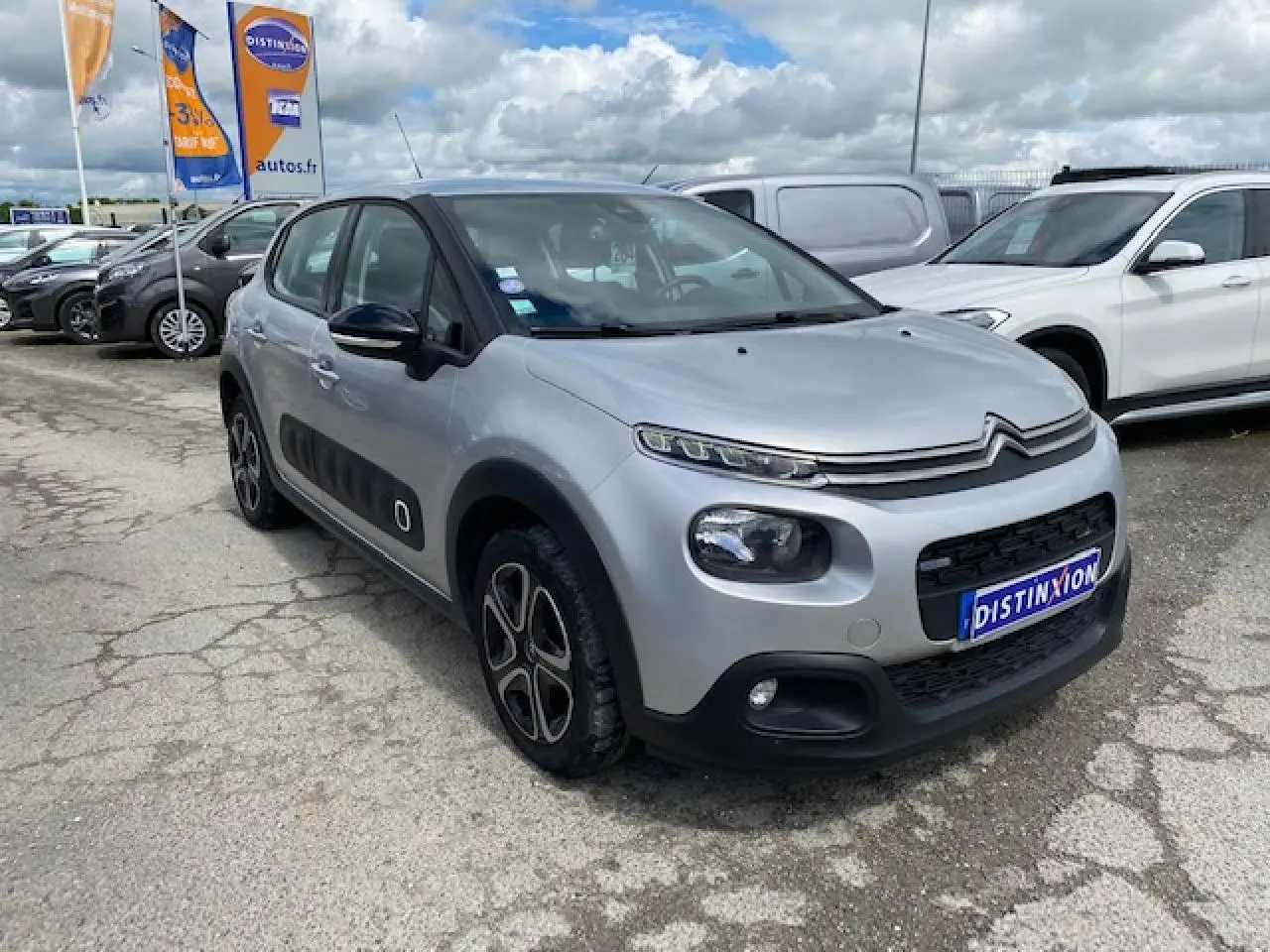 Citroën C3 gris clair vue 3/4 avant droit avec enjoliveurs 3D et protections latérales noires sur parking.
