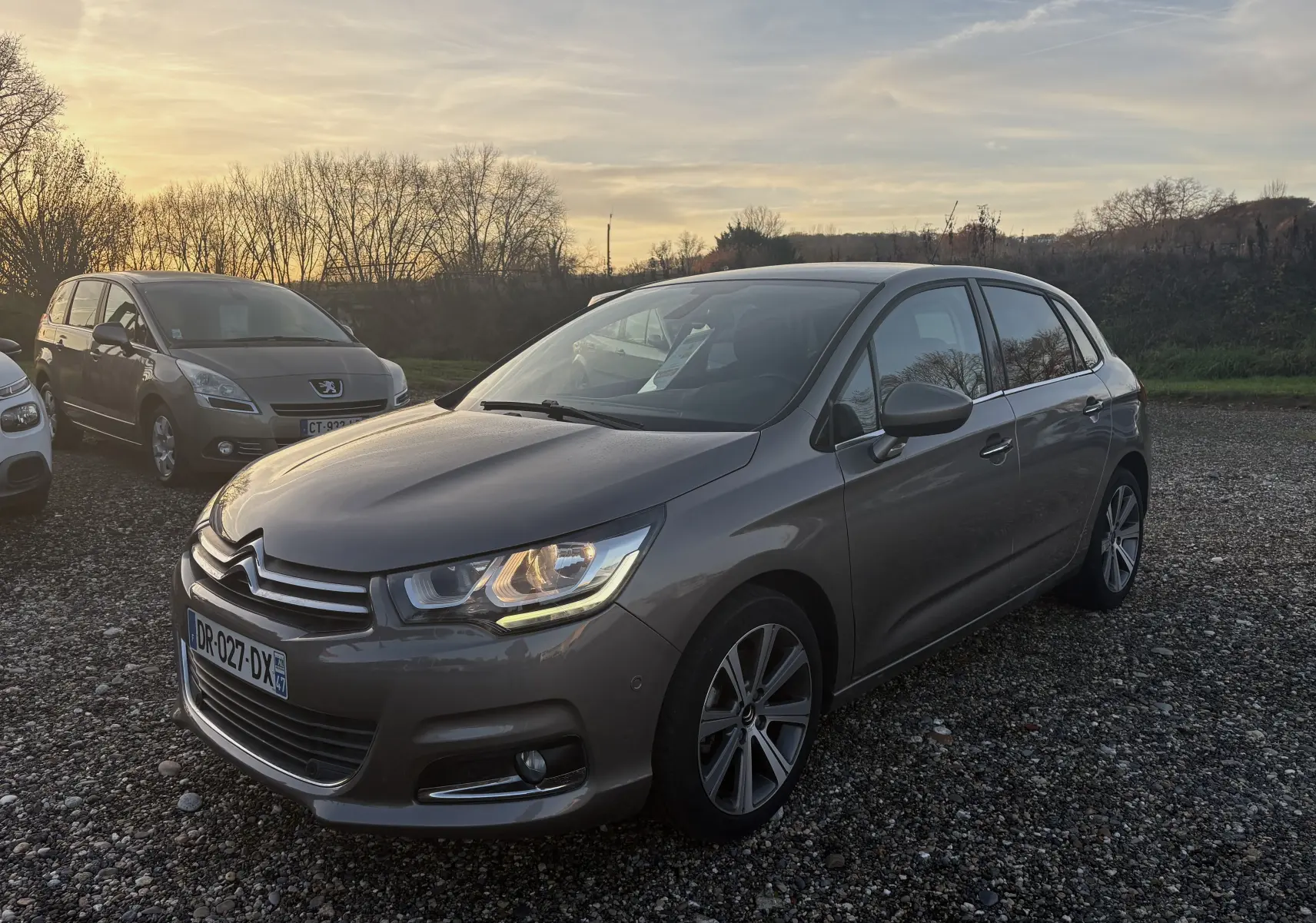 Citroën C4 II gris métallisé vue 3/4 avant droit au coucher du soleil sur un parking gravillonné.