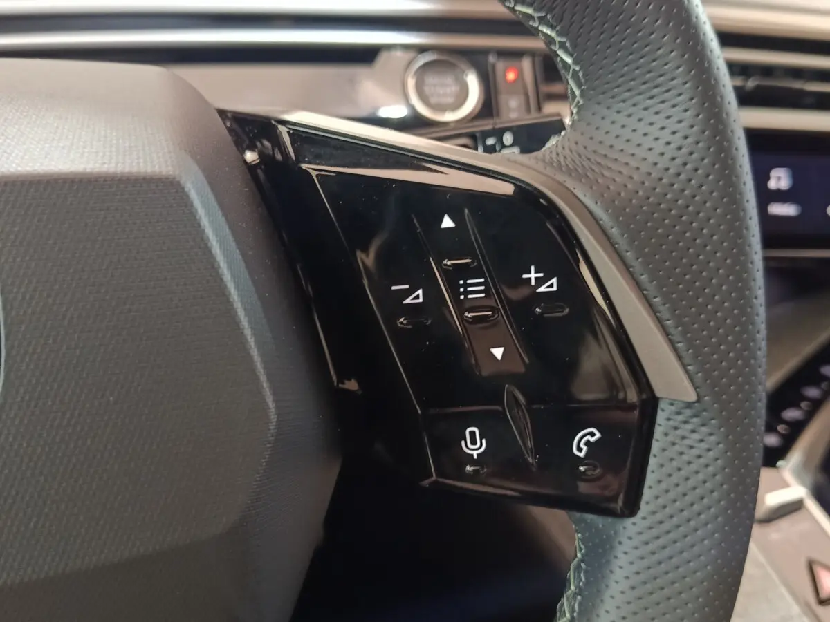 Gros plan sur les commandes noires au volant cuir perforé du Peugeot 5008 1.6 PHEV gris clair, intérieur moderne.