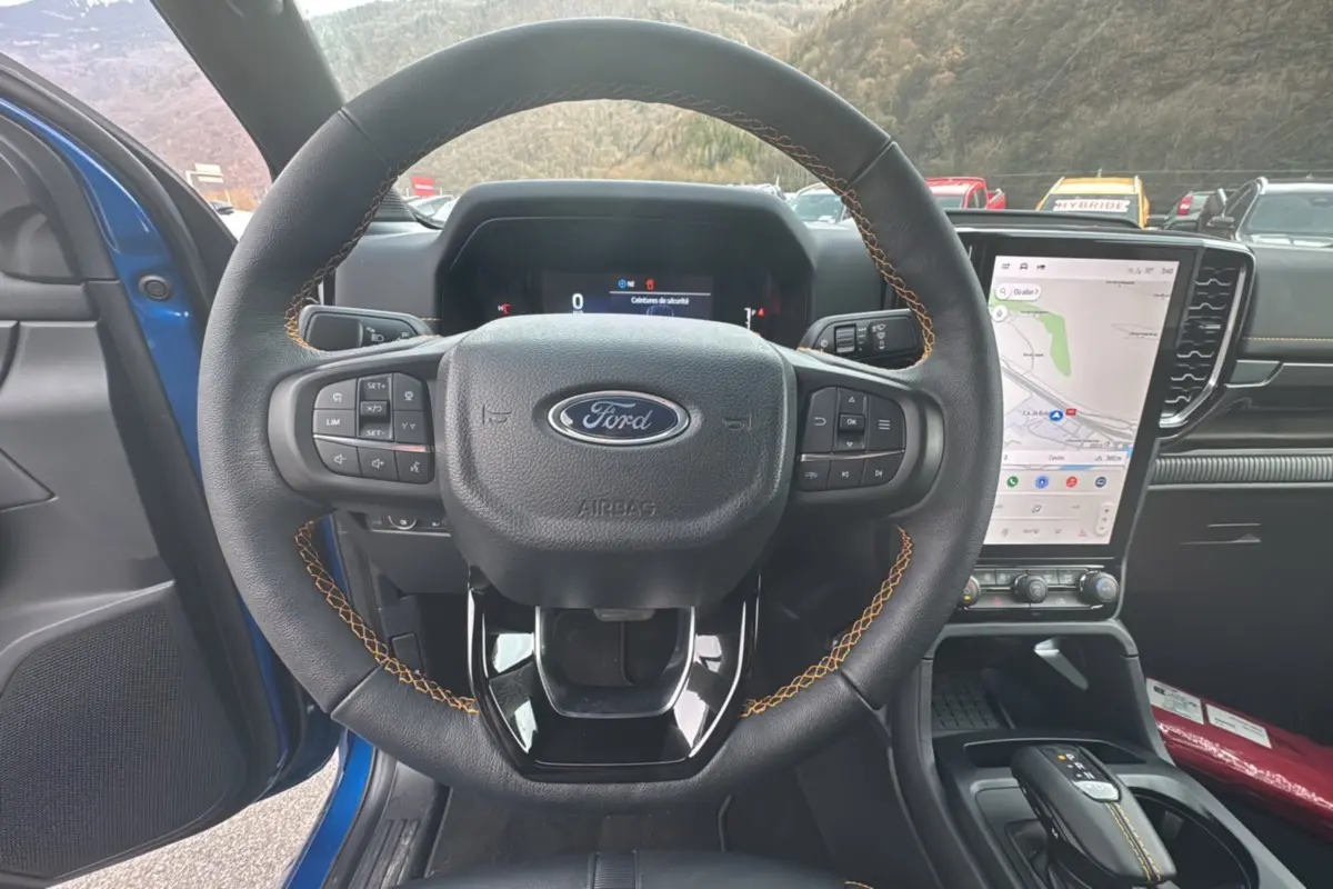 Vue centrée sur le volant cuir noir avec surpiqûres jaunes du Ford Ranger Wildtrak 2026, écran tactile vertical et intérieur bleu visible.