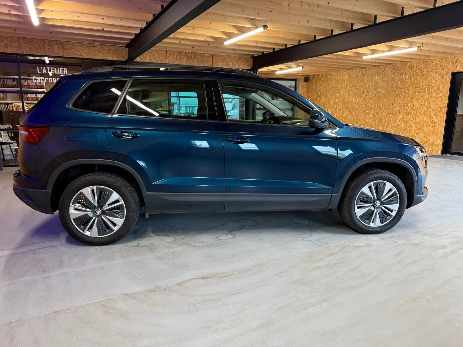 Vue de profil côté gauche d'un Skoda Karoq bleu 2023 avec jantes alu 17 pouces et toit noir dans un garage.