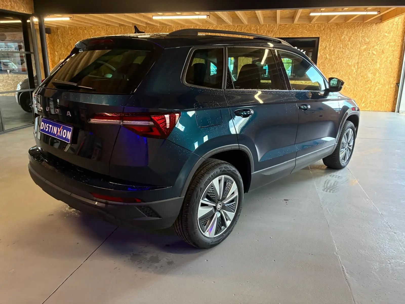 Vue 3/4 arrière droite d'un Skoda Karoq bleu 2023 avec jantes alu 17 pouces et feux arrière LED dans un garage.