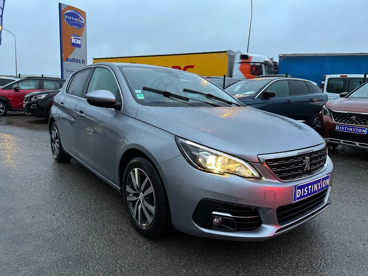 Peugeot 308 gris Artense vue 3/4 avant droit sous la pluie avec phares allumés sur parking extérieur.