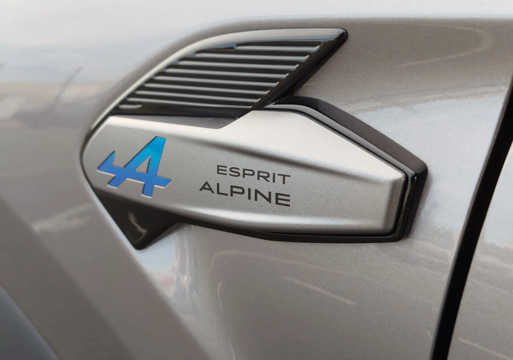 Gros plan sur l’insigne Esprit Alpine bleu et noir sur la carrosserie gris cassiopée métal d’une Renault Symbioz.