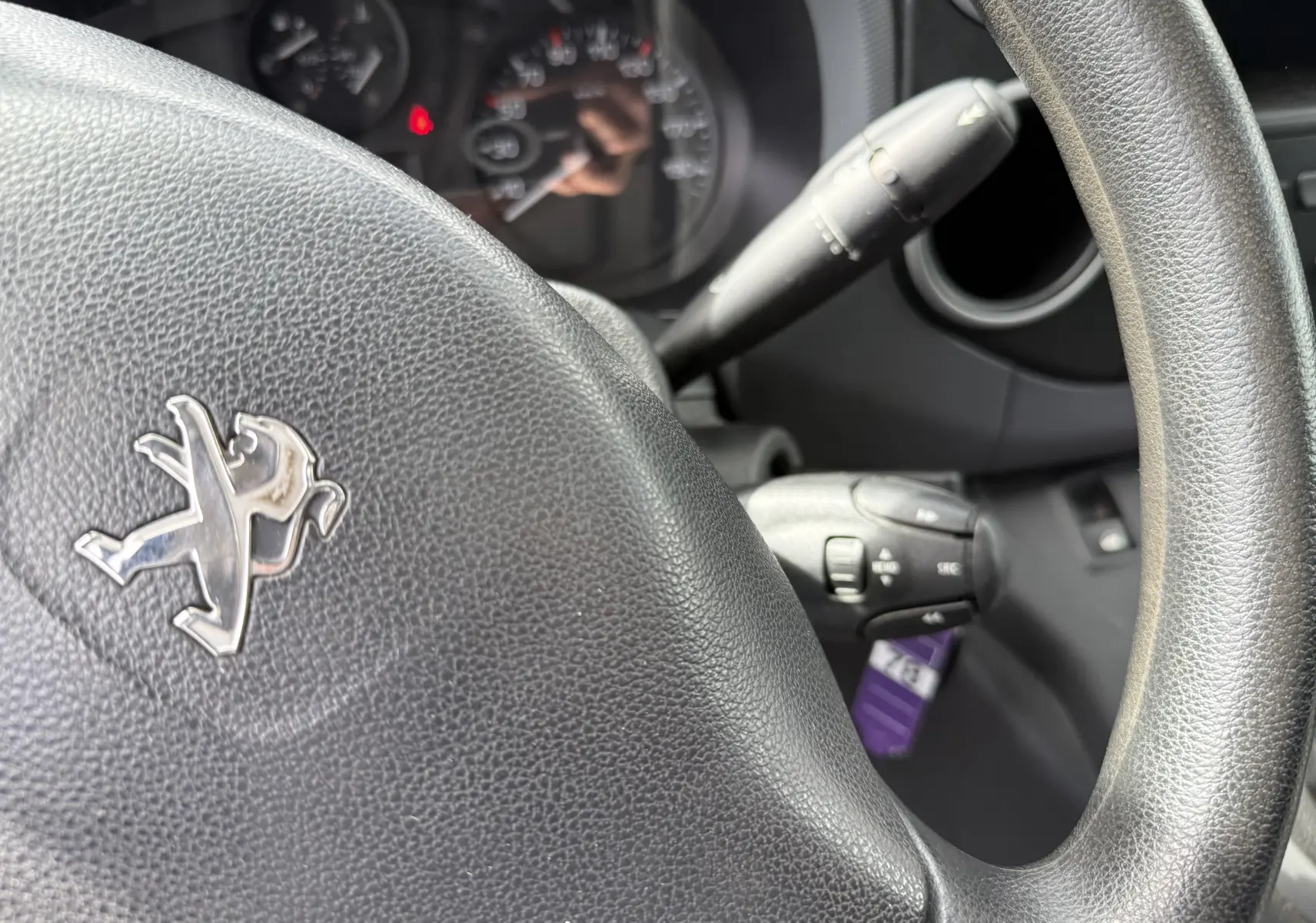 Gros plan sur le volant noir avec logo Peugeot du Partner 1.5 HDI 75, tableau de bord et commandes visibles en arrière-plan.