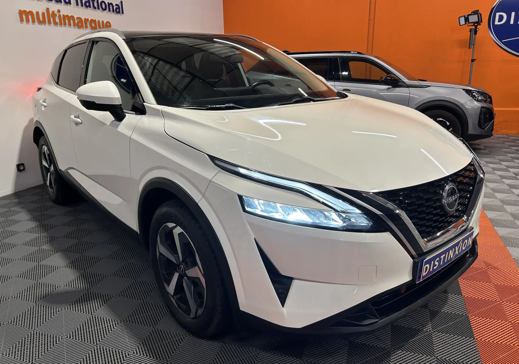 Nissan Qashqai blanc lunaire en 3/4 avant droit, avec phares LED allumés et toit panoramique visible.