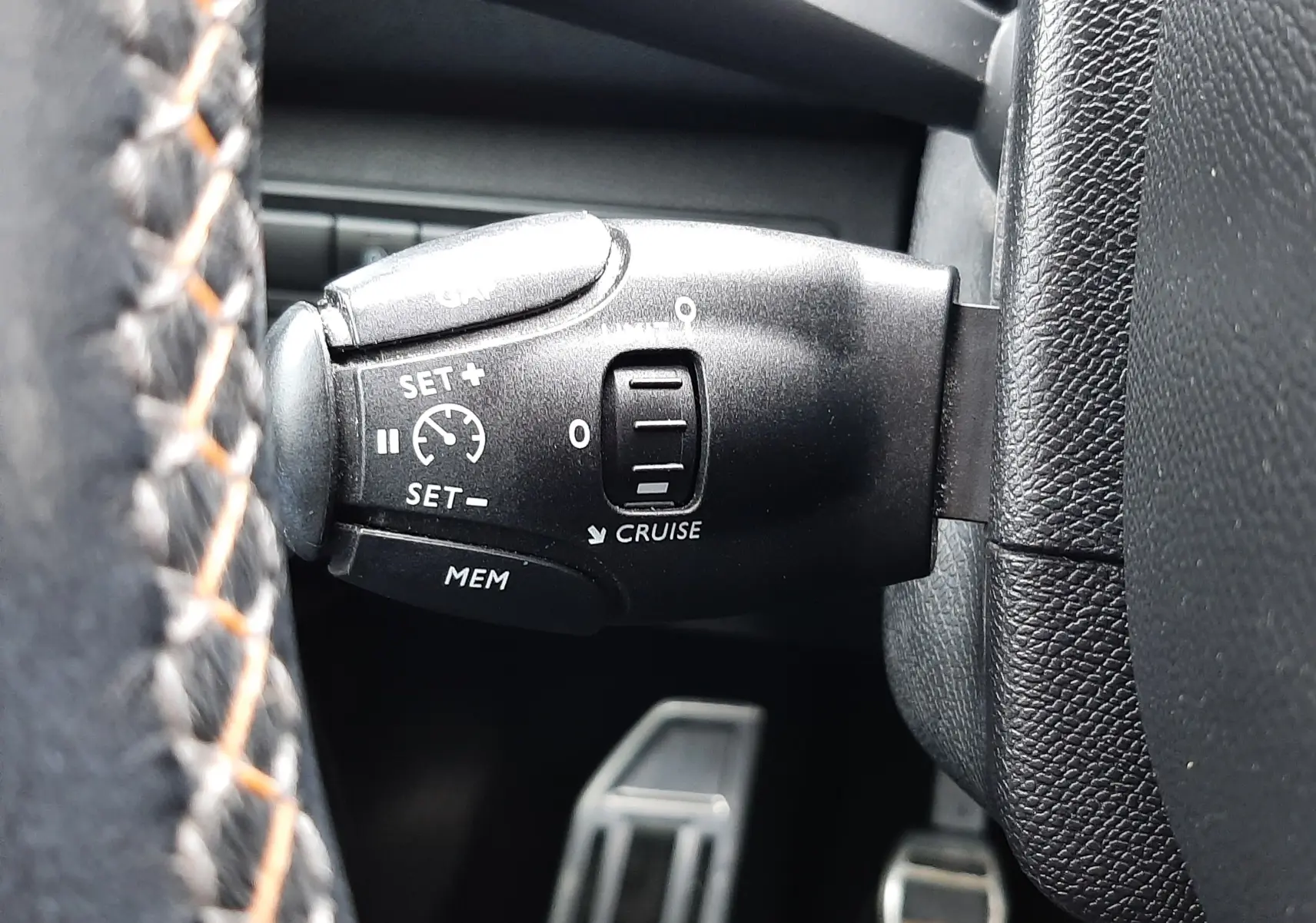 Gros plan sur la commande de régulateur de vitesse noire à gauche du volant, avec pédales en arrière-plan dans un intérieur noir.