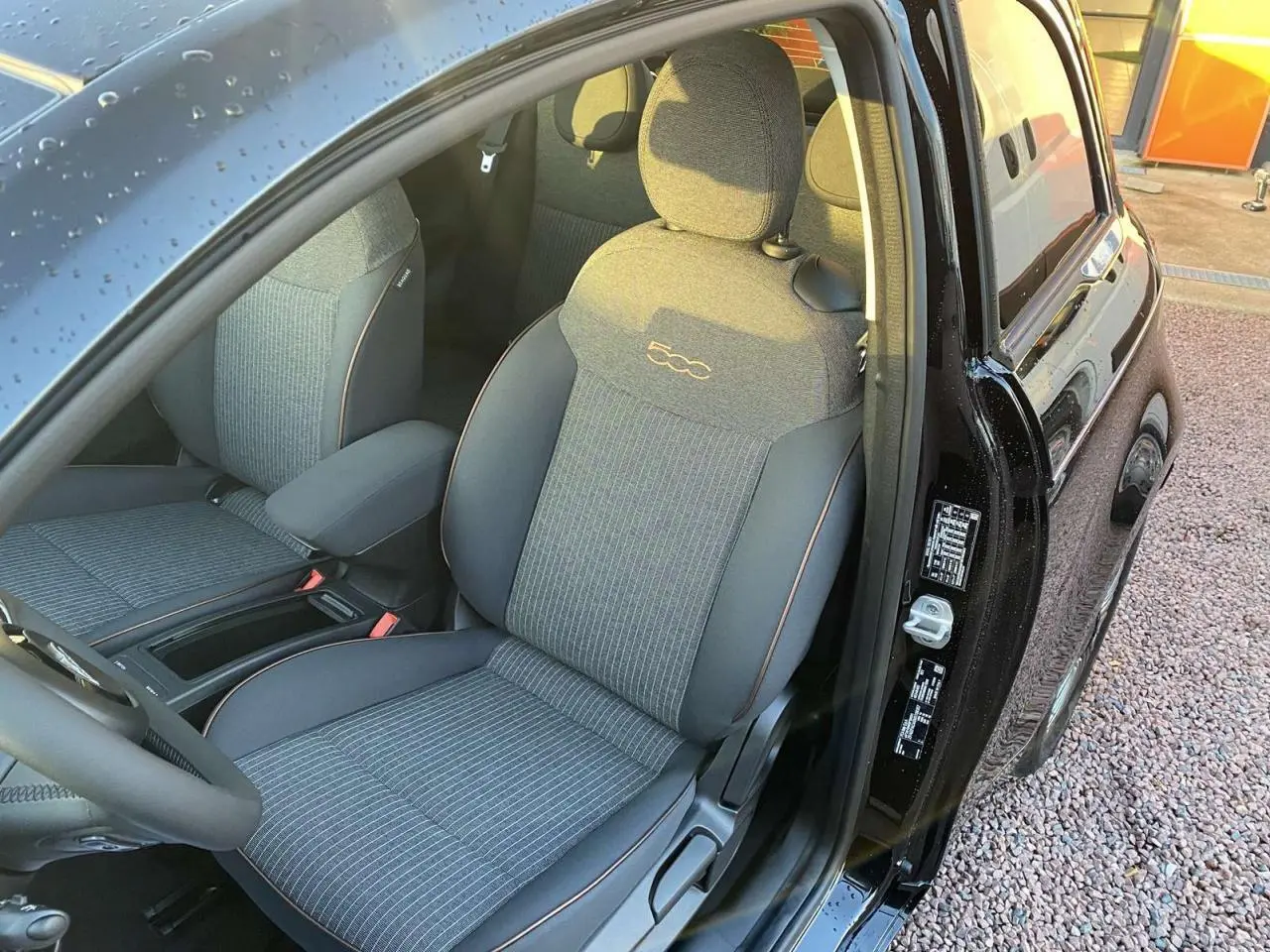 Vue intérieure côté conducteur d'une FIAT 500 électrique noire, siège avant gris avec logo 500 brodé et porte ouverte.