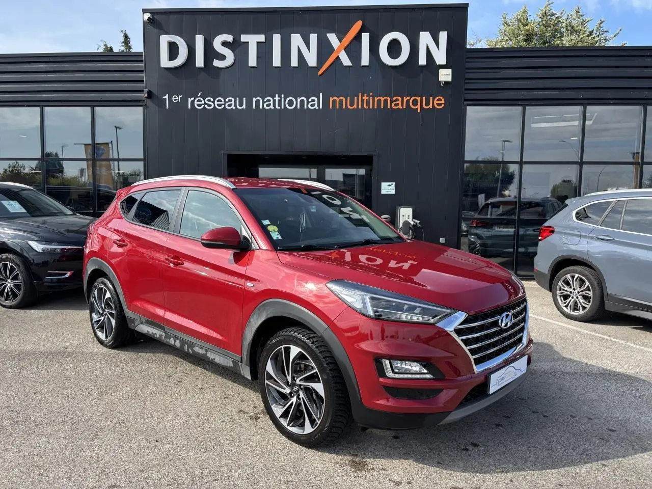 Hyundai Tucson rouge métallisé en 3/4 avant droit, avec jantes alliage et calandre chromée visible devant un bâtiment noir Distinxion.
