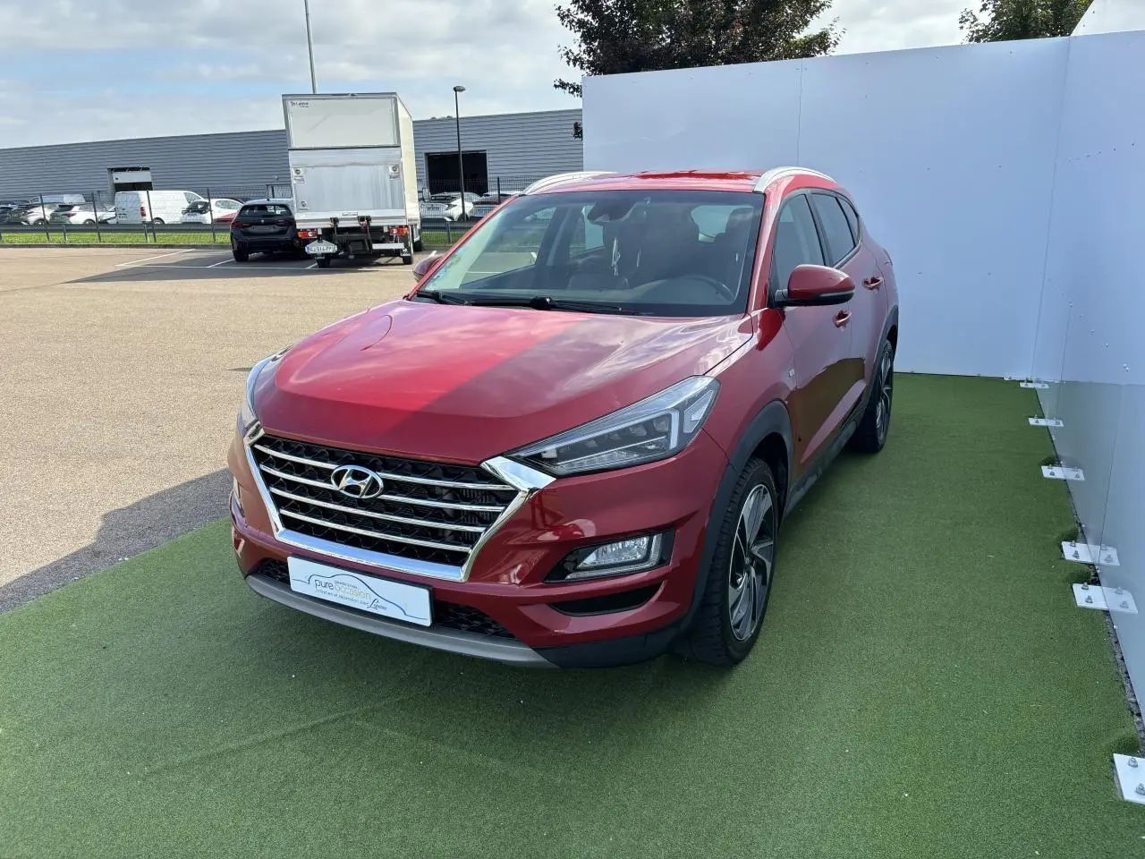 Vue 3/4 avant d'un Hyundai Tucson rouge métallisé garé sur un sol vert avec calandre chromée et jantes alliage.