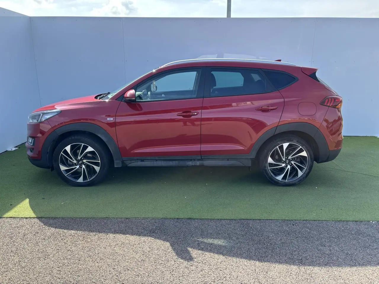 Profil côté gauche d'un Hyundai Tucson rouge métallisé avec jantes alliage et protections noires sur bas de caisse.