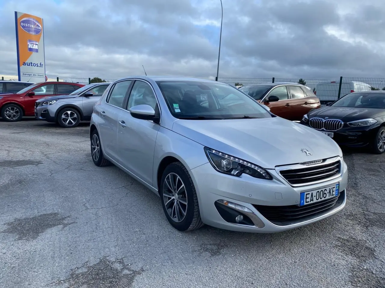 Peugeot 308 gris aluminium vue 3/4 avant droit, avec phares LED et jantes alliage sur parking extérieur.