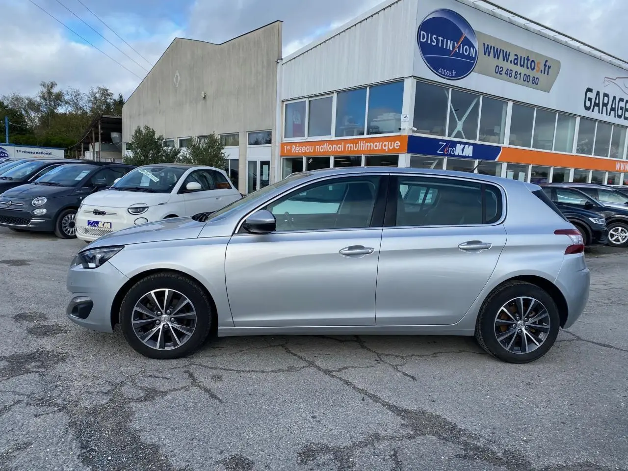 Profil côté gauche d'une Peugeot 308 gris aluminium 2016 garée devant un garage multimarque.