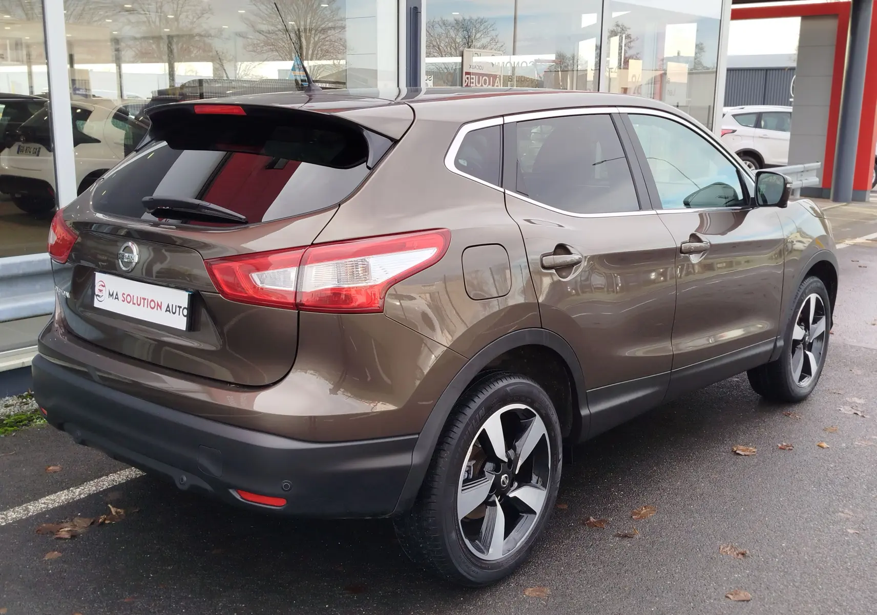 Vue 3/4 arrière droite d'un Nissan Qashqai bronze Ultime avec jantes alliage et vitres teintées, stationné devant un showroom.