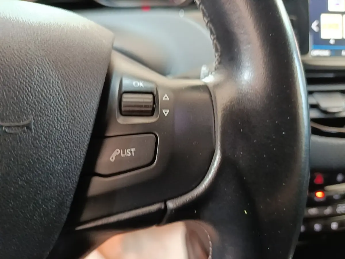 Gros plan sur le volant cuir noir de la Peugeot 2008 avec commandes vocales et écran tactile en arrière-plan flou.