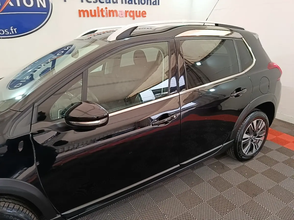 Vue côté gauche d'un Peugeot 2008 noir avec jantes alliage et barres de toit chromées en intérieur showroom.