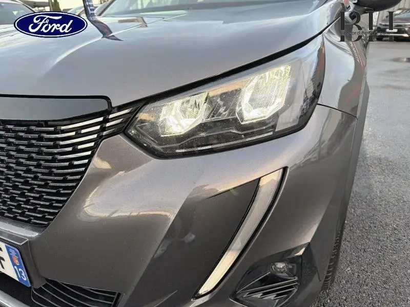 Gros plan sur le phare avant droit et la calandre d'une Peugeot 2008 gris Artense avec feux LED allumés.