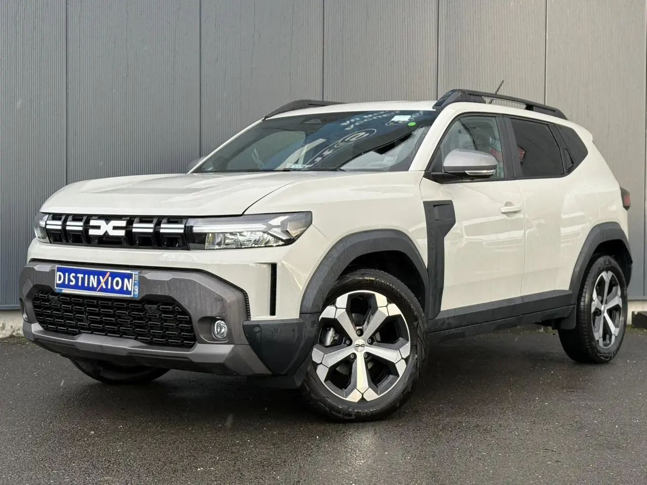 Vue 3/4 avant droite du Dacia Duster 2024 beige Sandstone avec barres de toit noires et jantes alliage 18 pouces.