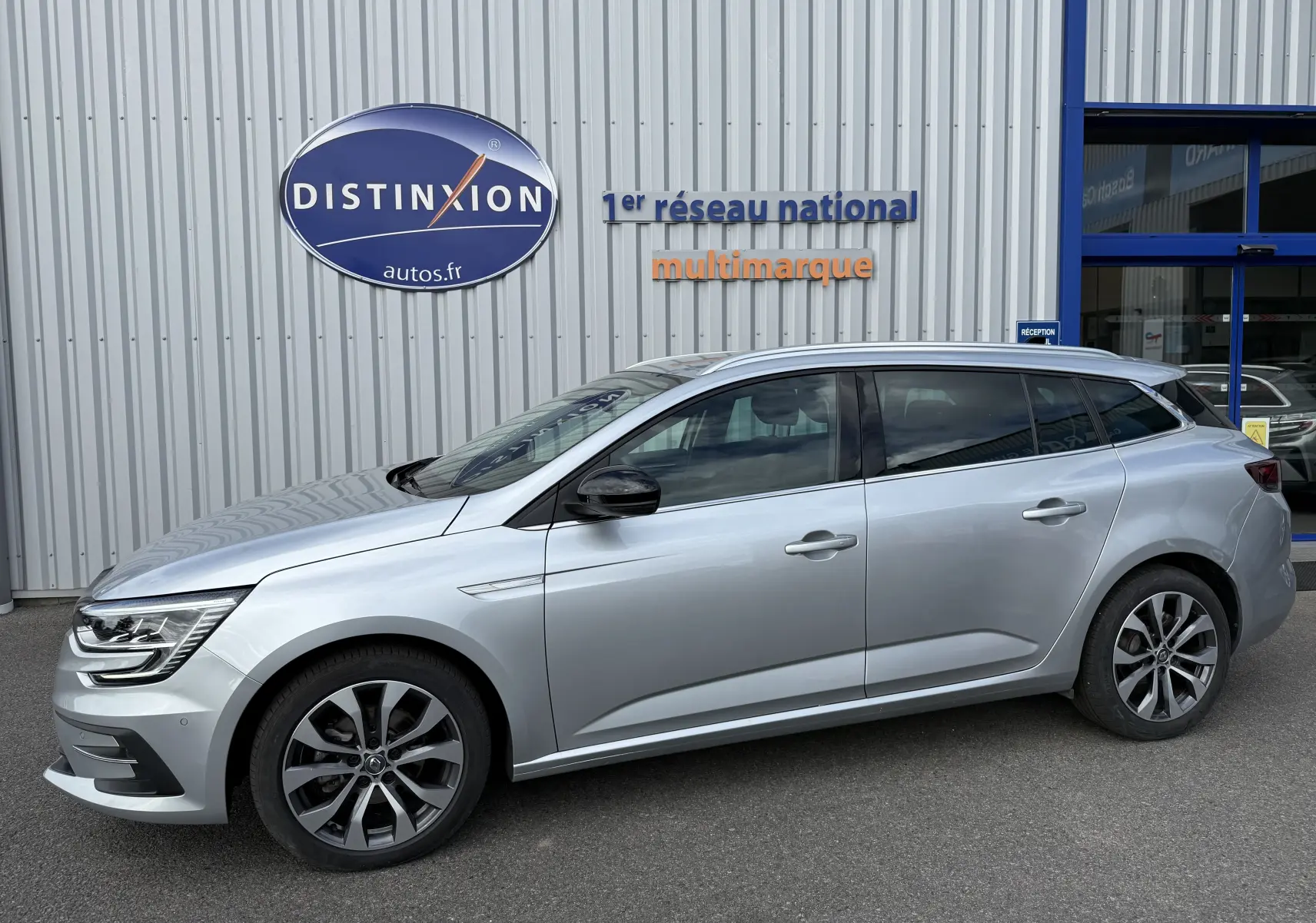 Profil droit d'une Renault Mégane Break gris Highland Metal 2024 stationnée devant un bâtiment commercial.