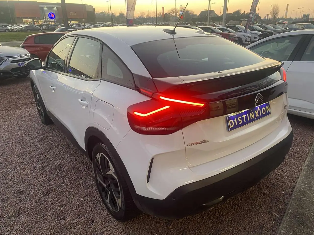 Vue 3/4 arrière droite d'une Citroën C4 blanche avec feux arrière allumés sur parking en soirée.