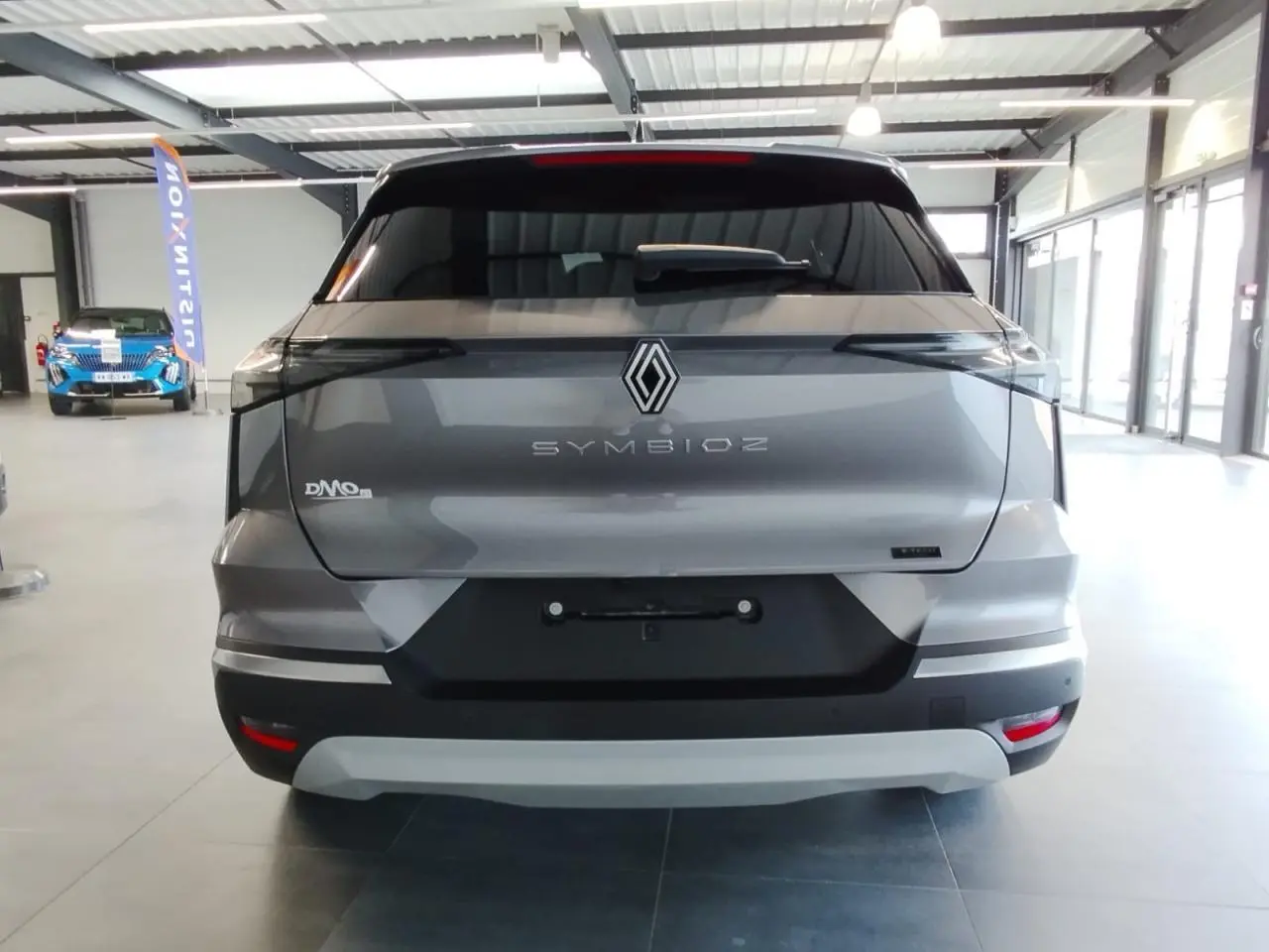 Vue arrière d’un Renault Symbioz gris Cassiopée avec logo central et feux arrière noirs étirés en intérieur showroom.