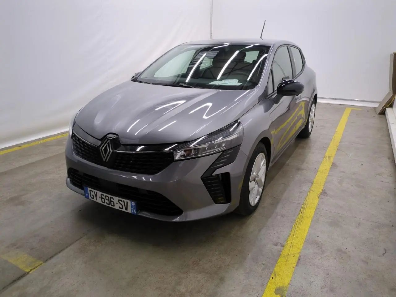 Renault Clio 2024 gris foncé vue 3/4 avant droit dans un garage avec marquages au sol jaunes.