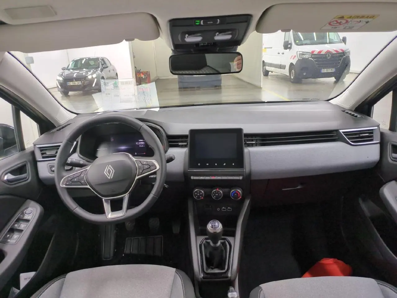 Vue intérieure avant de la Renault Clio 2024 avec tableau de bord gris, volant multifonction et écran tactile central.