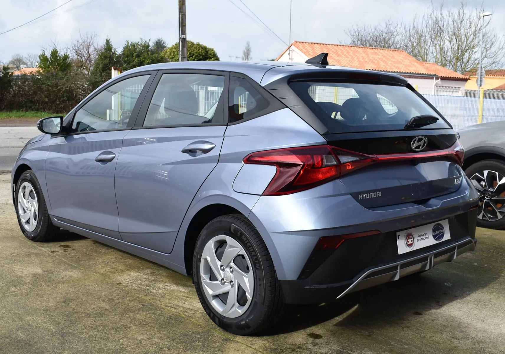 Vue 3/4 arrière gauche d'une Hyundai i20 bleu clair 2025 avec spoiler de toit et feux arrière en bandeau lumineux.