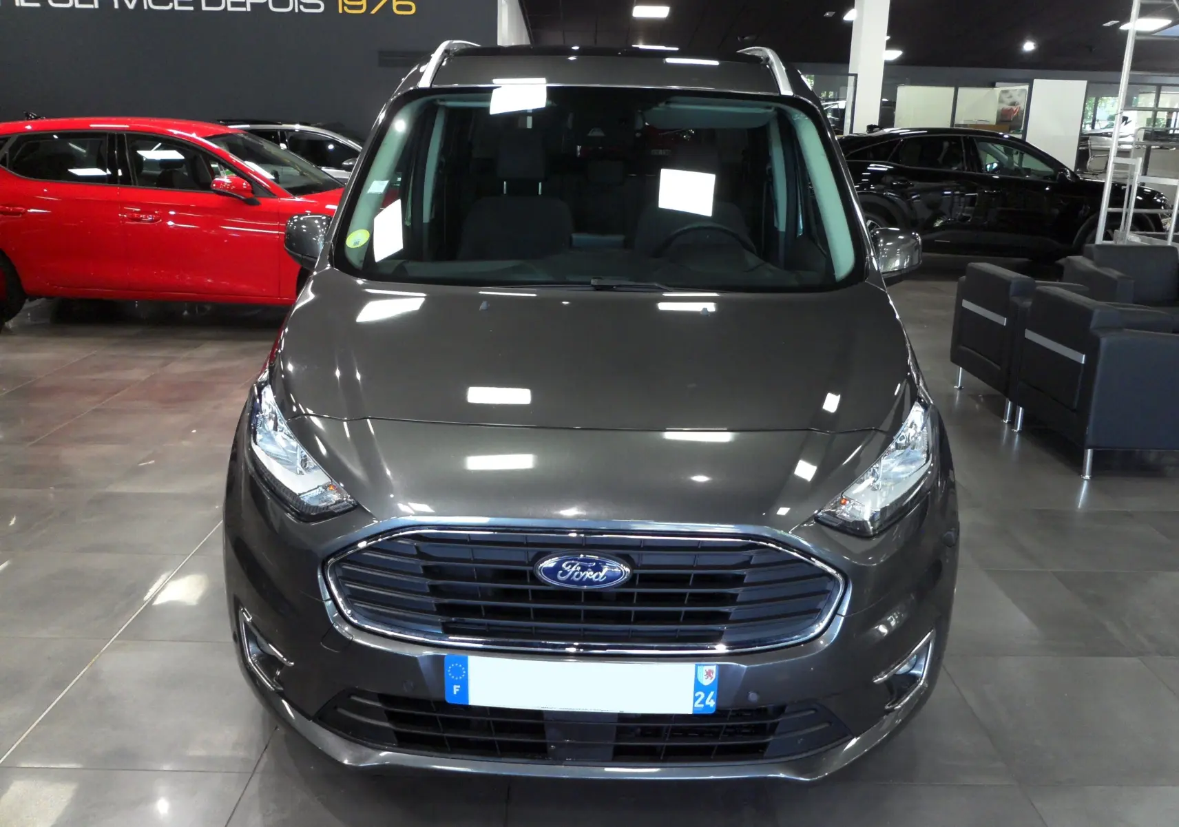 Vue frontale d'un Ford Grand Tourneo Connect gris Magnetic exposé en showroom avec calandre chromée et phares allumés.
