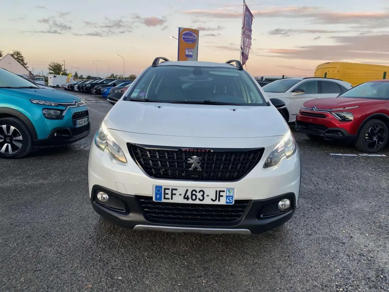 Peugeot 2008 blanc vue de face, avec calandre noire et phares allumés sur un parking extérieur.
