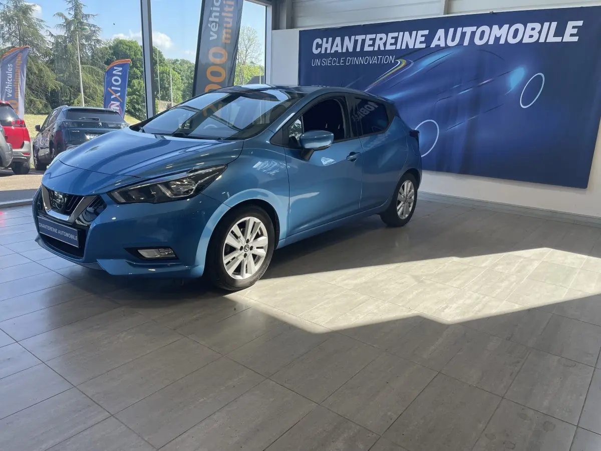 Nissan Micra IG-T 100 N-Connecta bleu vue 3/4 avant droit en showroom avec éclairage naturel et logo concession visible.