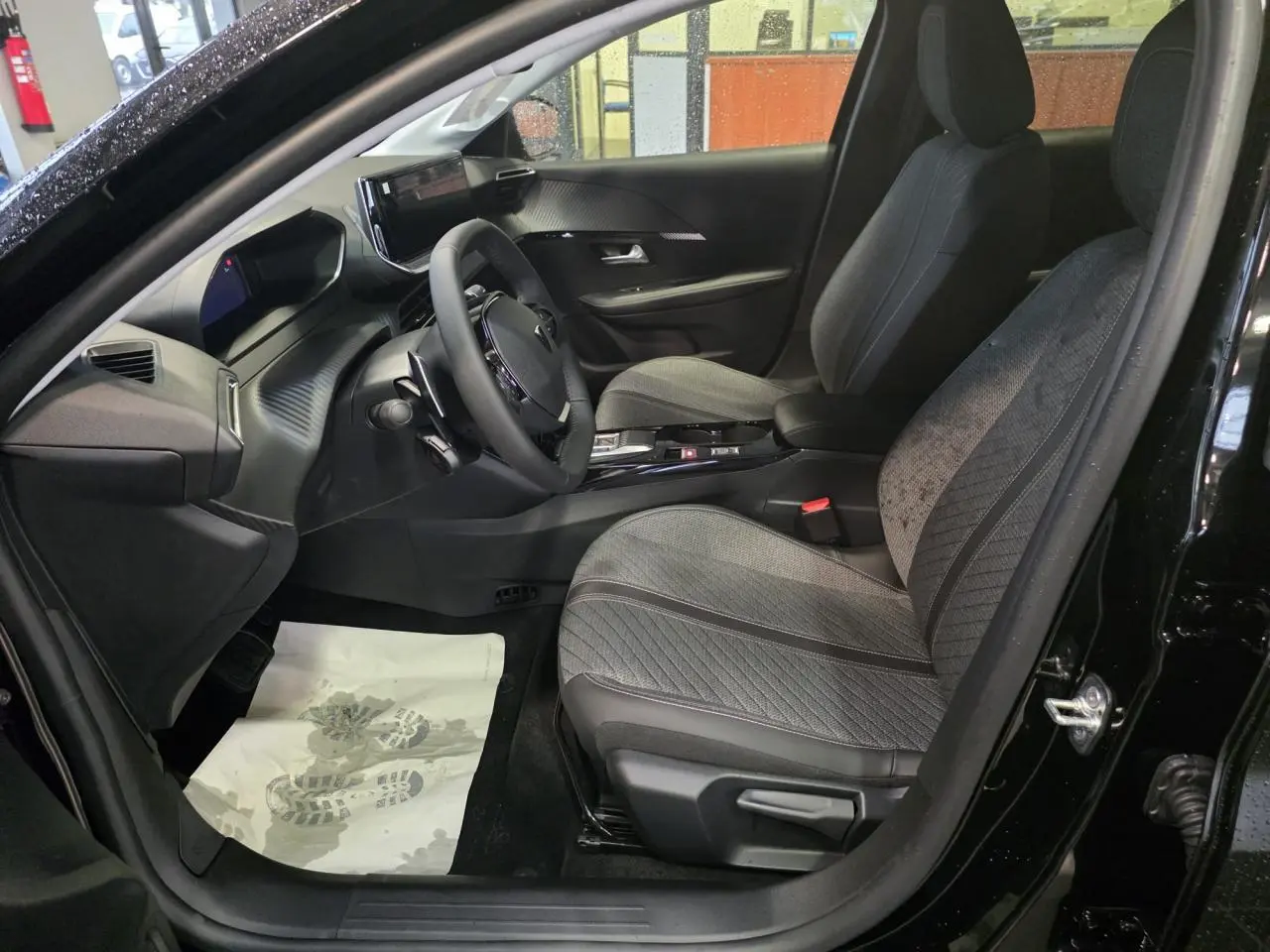 Vue intérieure côté conducteur de la Peugeot 208 noire métallisée, avec sièges tissu gris et tableau de bord moderne.