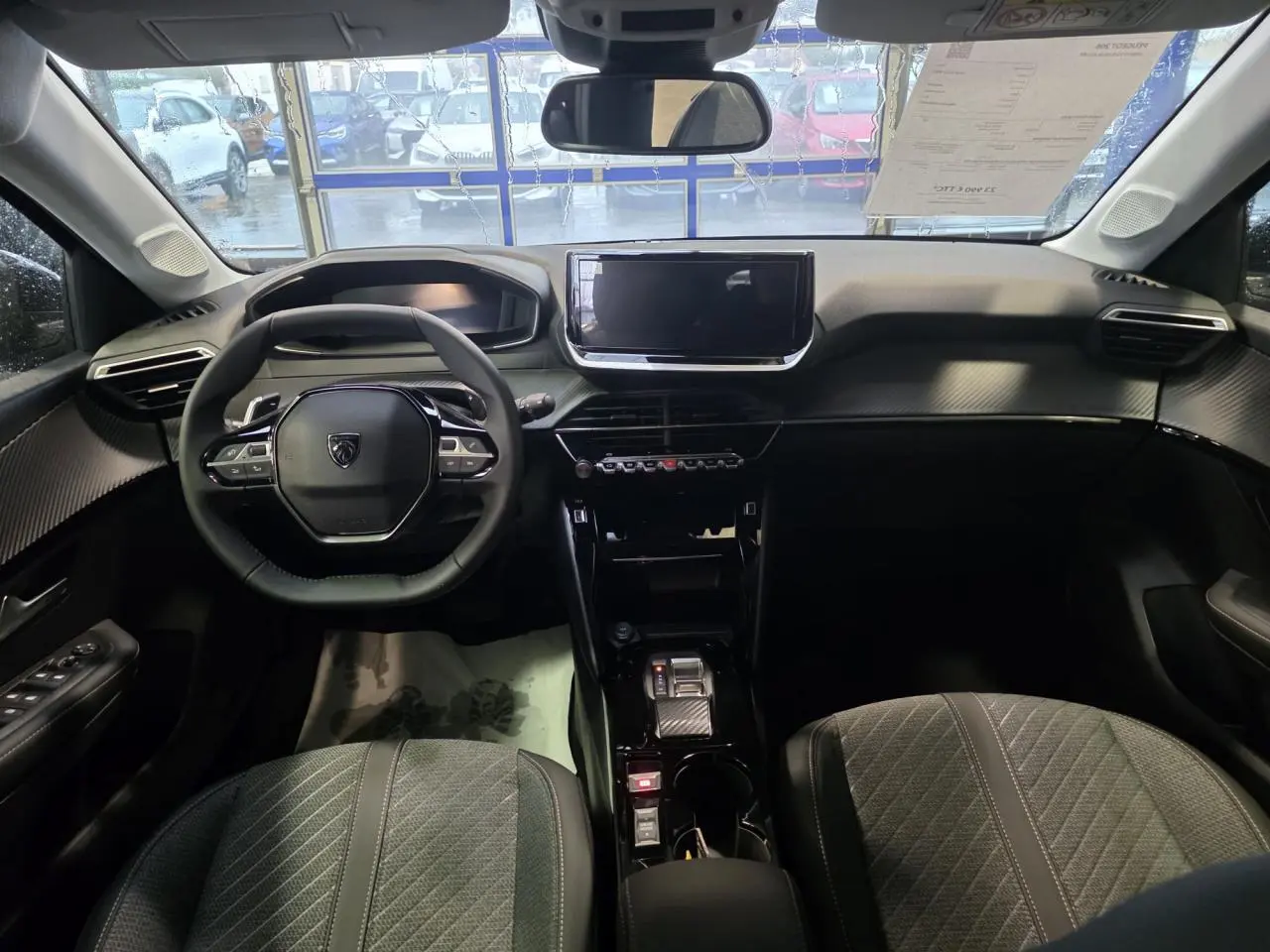 Vue intérieure avant de la Peugeot 208 1.2i Hybrid 2025, tableau de bord noir avec volant compact et écran tactile central.