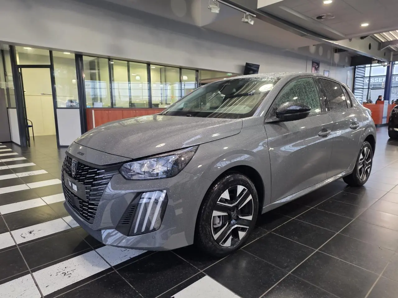 Peugeot 208 gris selenium en 3/4 avant droit, avec calandre noire et jantes alliage bi-ton.