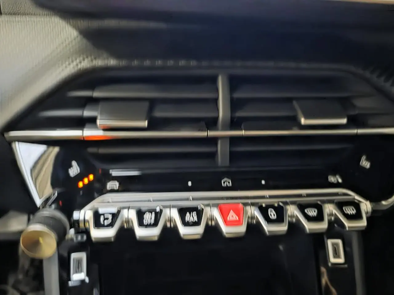 Détail de la console centrale noire brillante de la Peugeot 208 1.2i Hybrid avec boutons argentés et bouton d’alerte rouge.