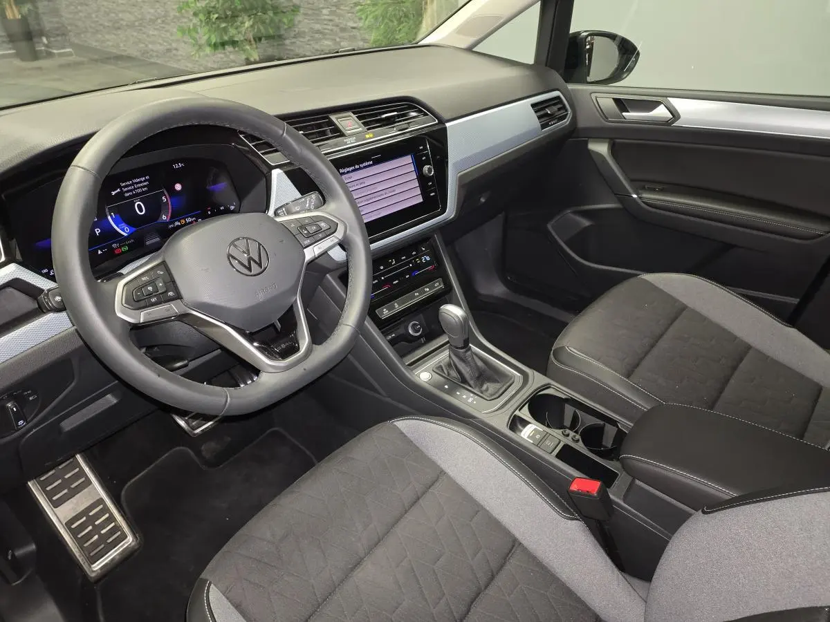 Vue intérieure côté conducteur du Volkswagen Touran 2025, montrant le volant cuir multifonction et la console centrale noire.