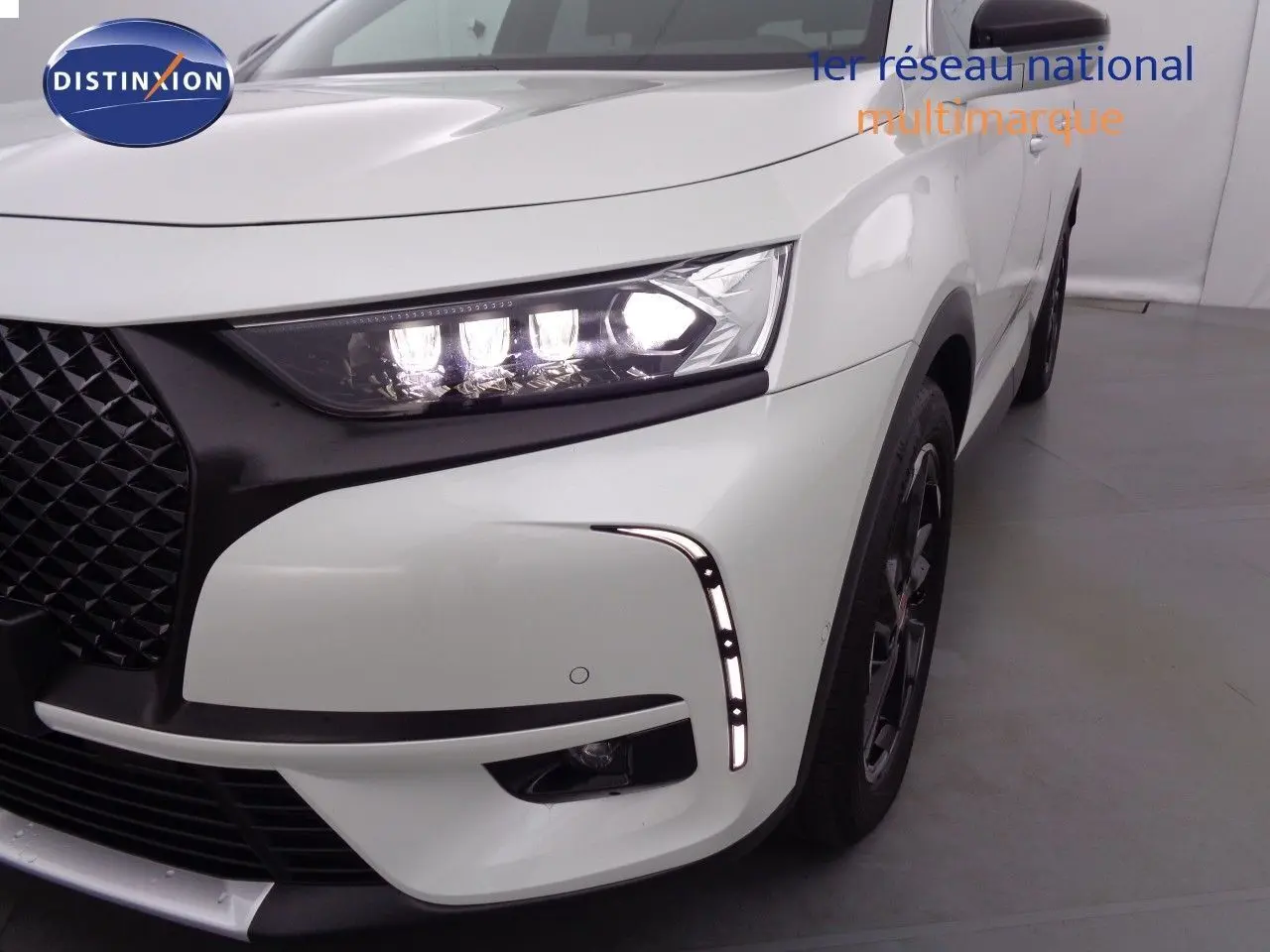Gros plan sur l'avant droit du DS7 Crossback blanc nacré, mettant en valeur le phare LED allumé et la calandre noire texturée.