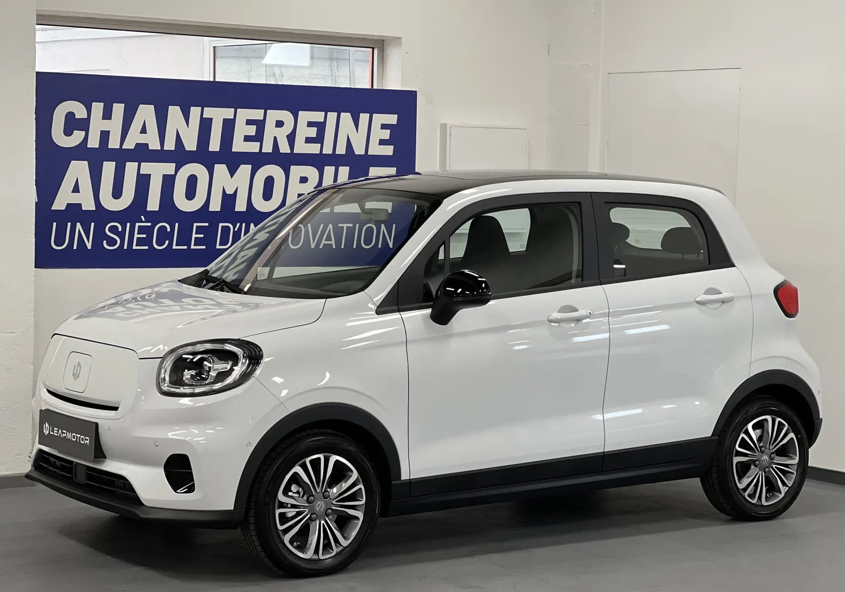 Vue 3/4 avant droite d'une Dacia Spring blanche électrique avec toit noir et jantes alliage en intérieur showroom.