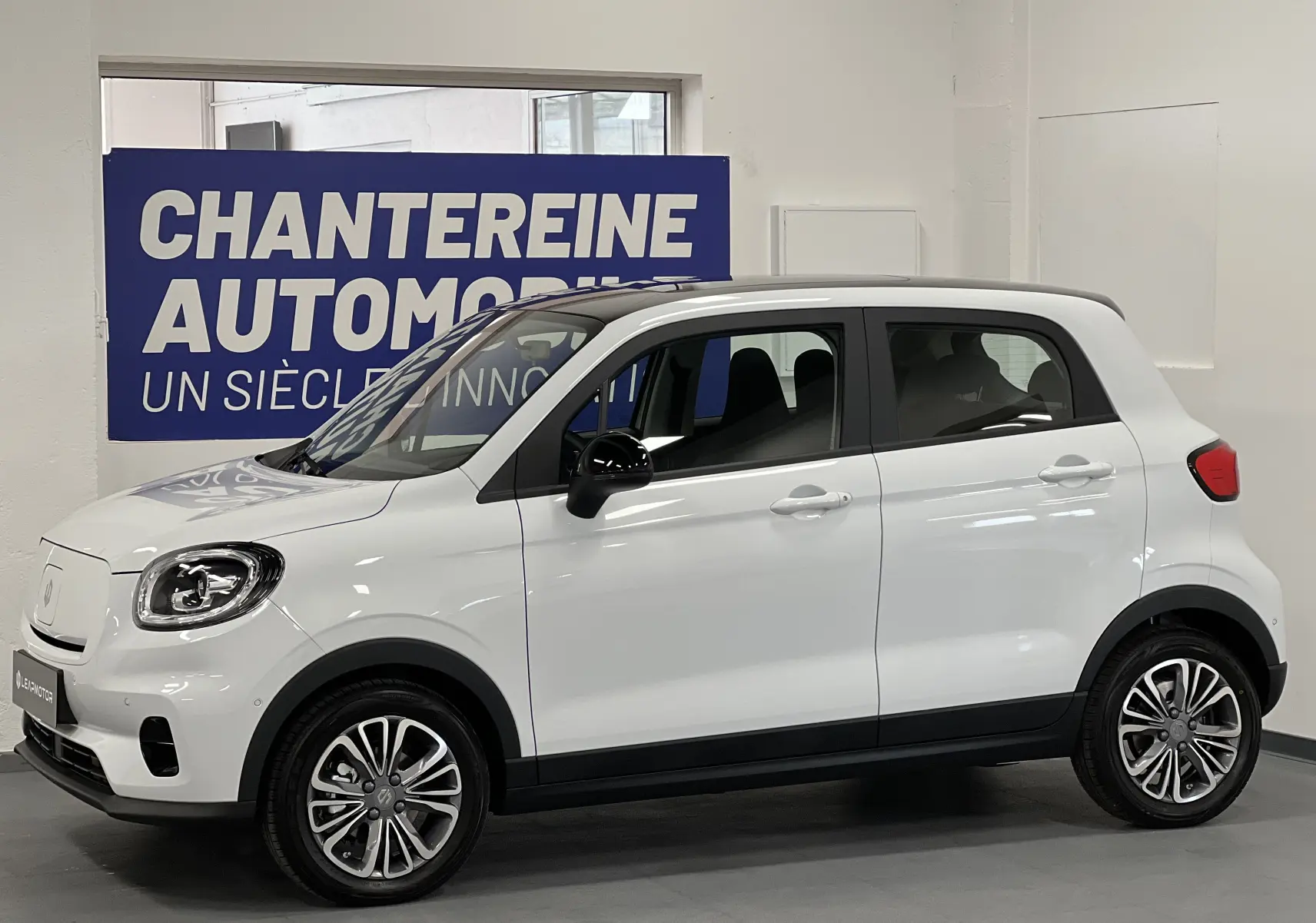 Vue de profil côté gauche d'une Dacia Spring blanche électrique avec jantes alliage et rétroviseurs noirs en intérieur showroom.