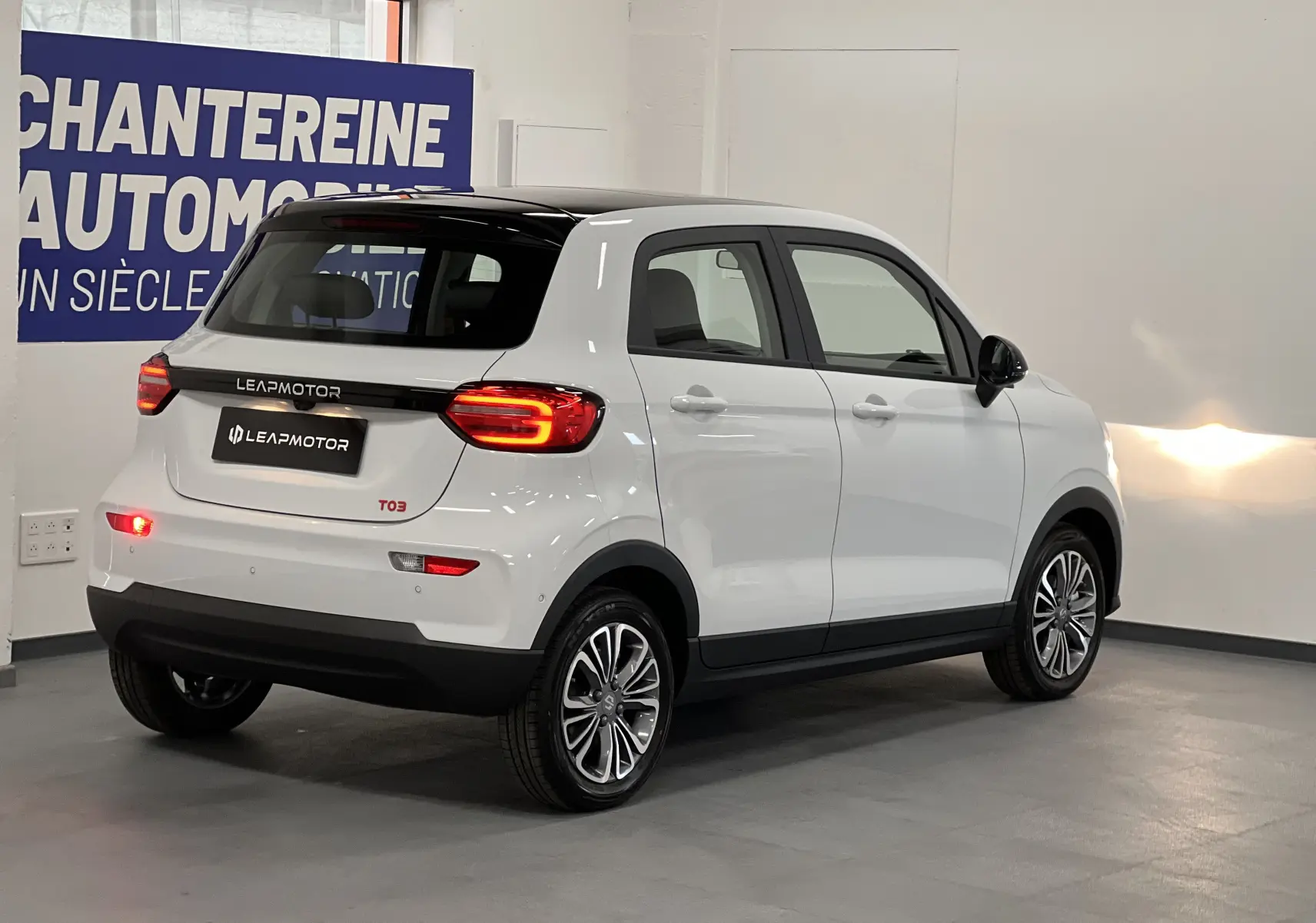 Vue 3/4 arrière droite d'une Dacia Spring blanche électrique avec toit noir et feux arrière allumés en intérieur showroom.