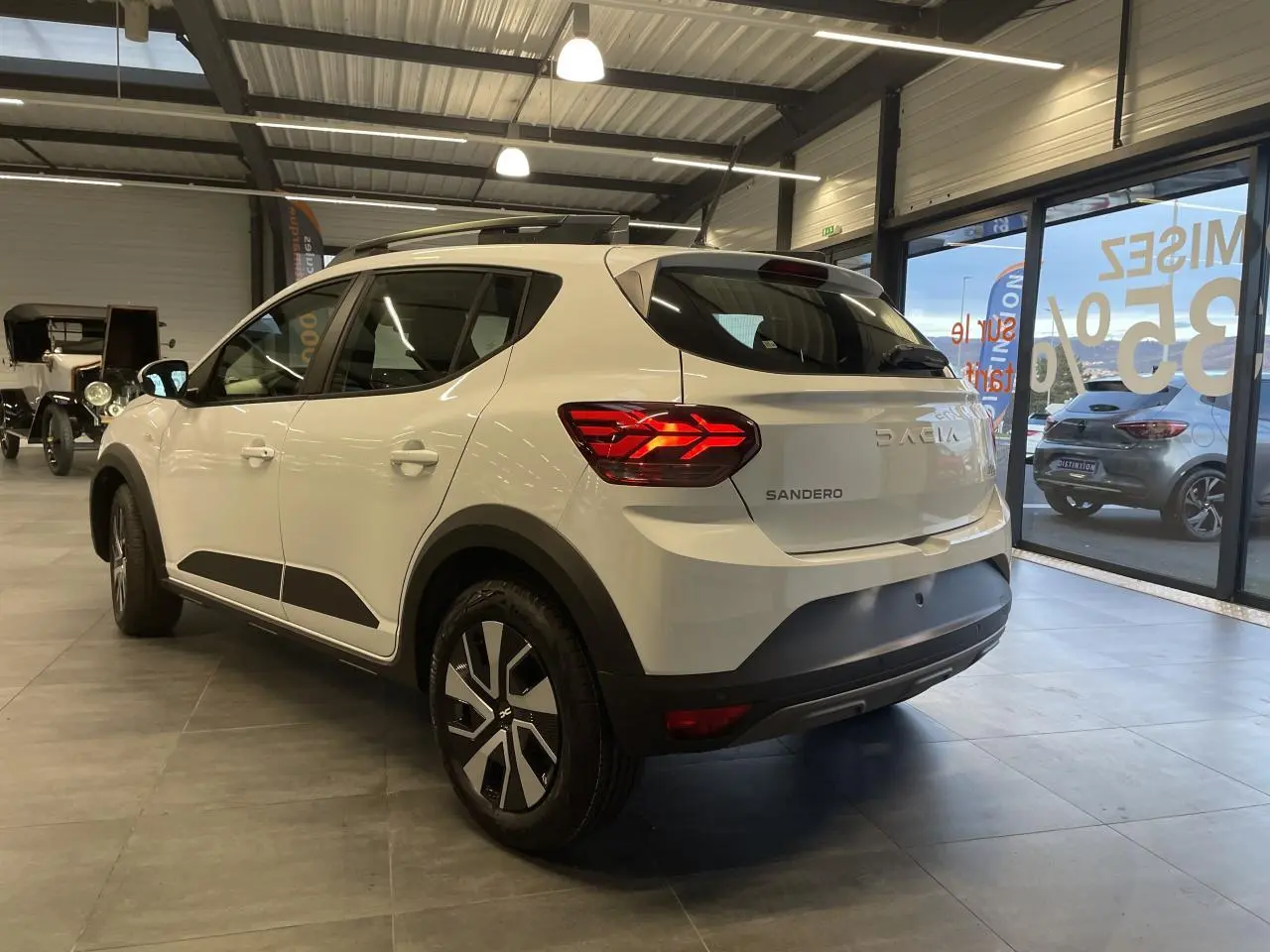 Vue 3/4 arrière droite d'une Dacia Sandero Stepway blanche Glacier avec barres de toit et feux arrière LED en intérieur showroom.