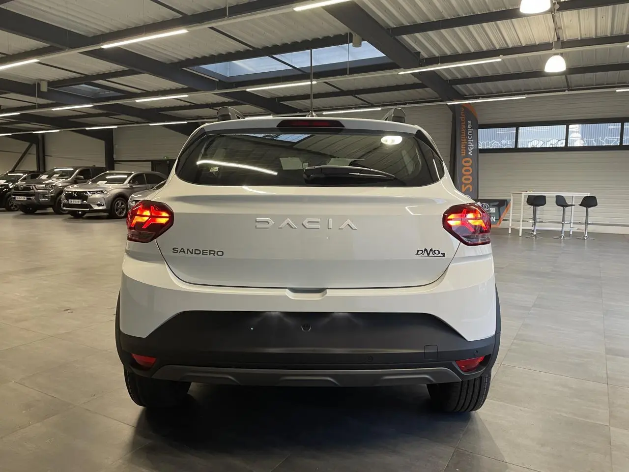 Vue arrière d'une Dacia Sandero TCe 90 Stepway blanche Glacier avec feux allumés dans un showroom.