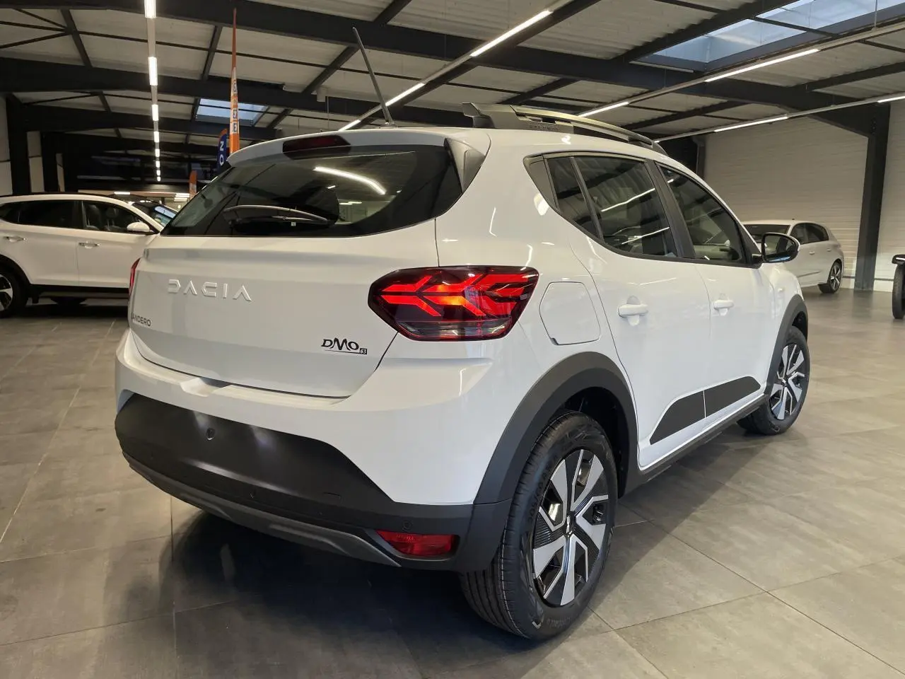 Vue 3/4 arrière droite d'une Dacia Sandero Stepway blanche avec feux arrière LED et barres de toit gris Mégalithe.