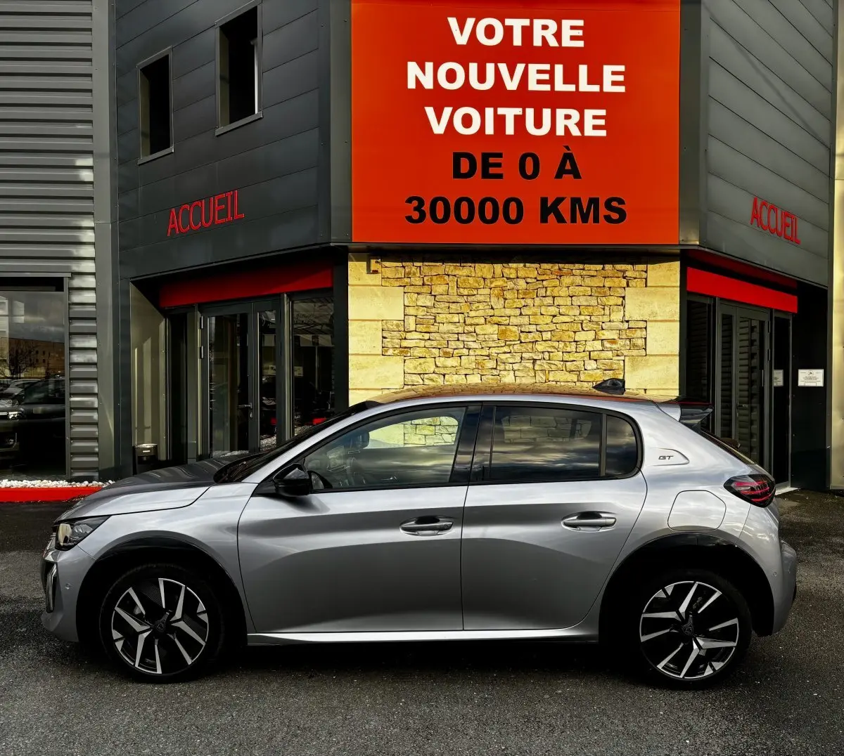 Peugeot 208 Hybrid 110 e-DCS6 GT gris clair vue de profil côté gauche devant un bâtiment avec panneau orange.